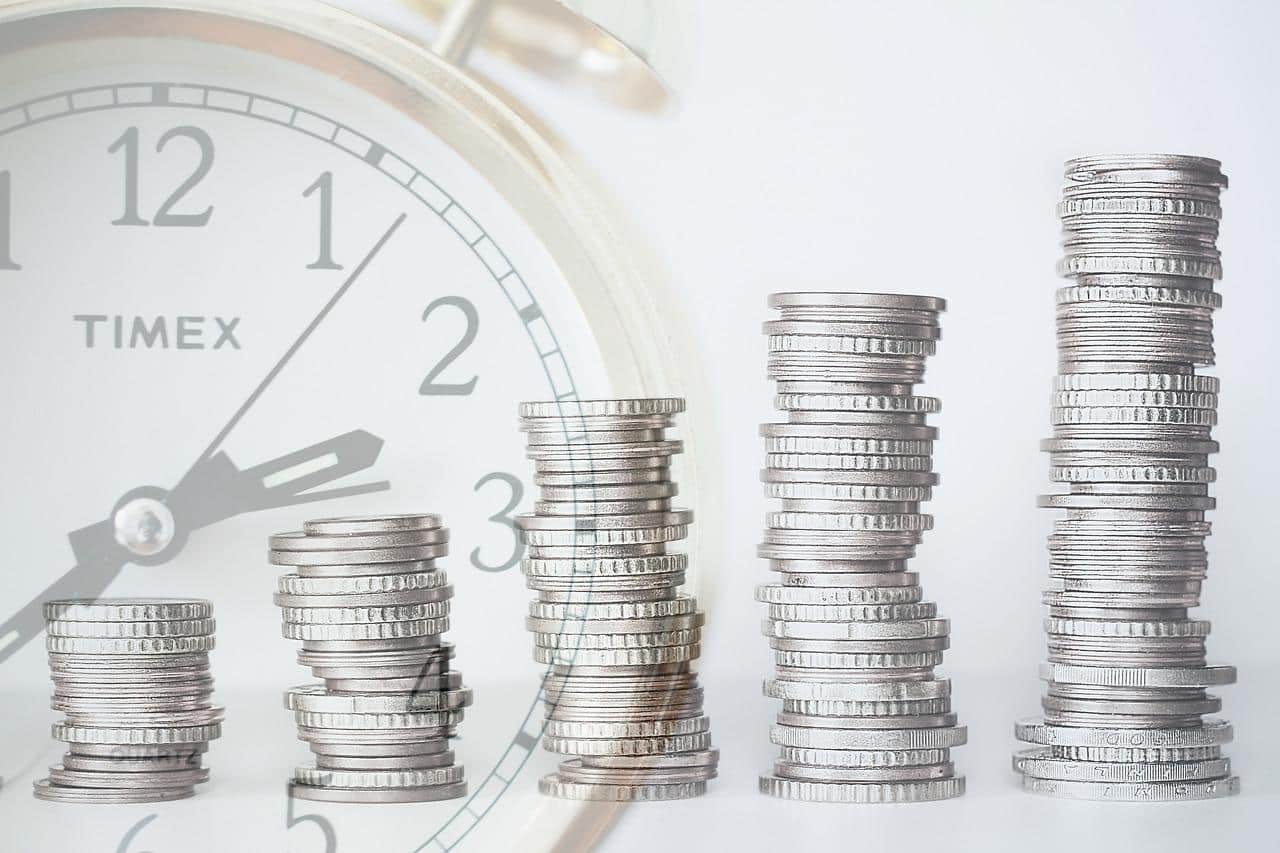 Stacks of coins getting larger moving left to right with a clock in the background. Meant to symbolize money growing over time or a positive return on investment