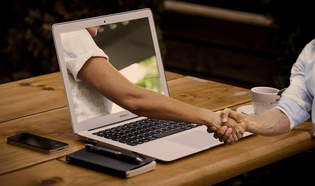 A hand reaching out of a laptop screen to shake another person's hand.