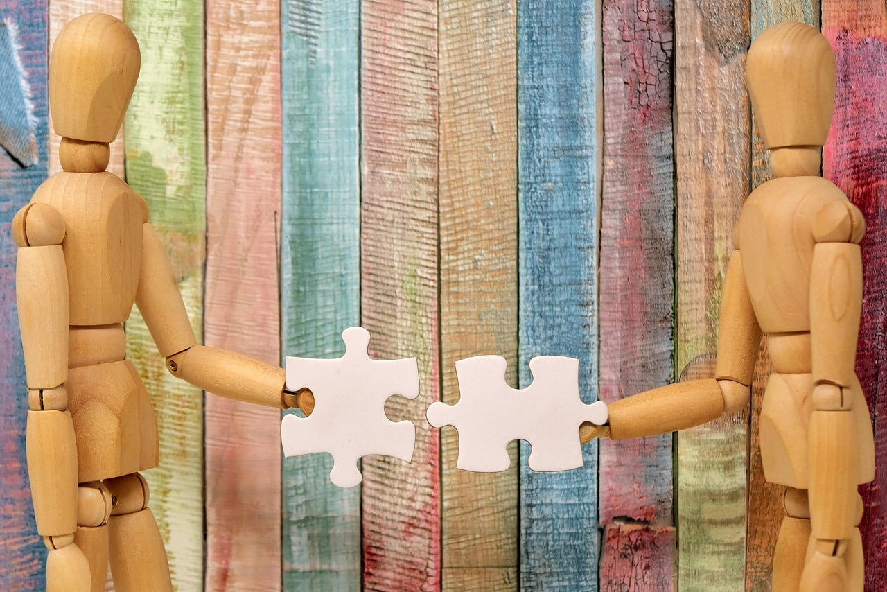 An image of two wooden figures holding large puzzle pieces and approaching one another as if they are going to connect them. This is in front of a fence board colored in different colors. Image meant to signify culture fit.