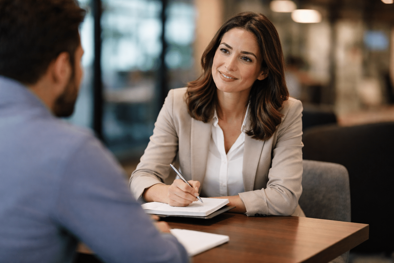 A professional woman in a beige blazer attentively listening to a client across a table in a modern office, holding a pen and notebook, with a warm, engaged expression.