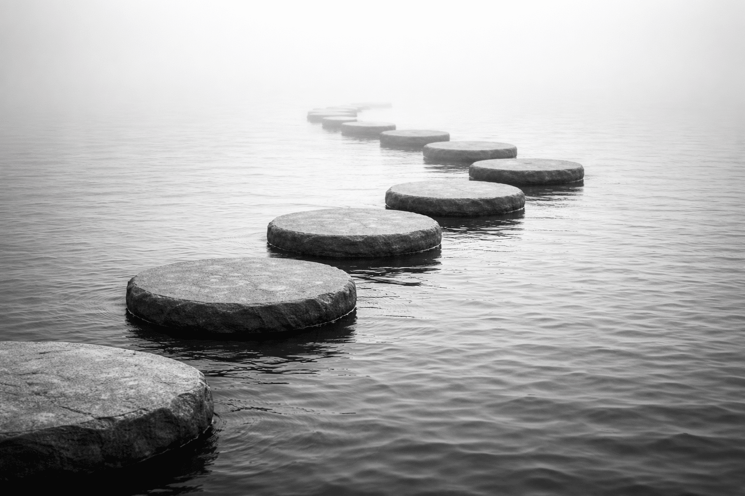 Black-and-white image of stepping stones forming an S-shaped path across calm water, with the first and last stones partially visible, suggesting continuous progress.